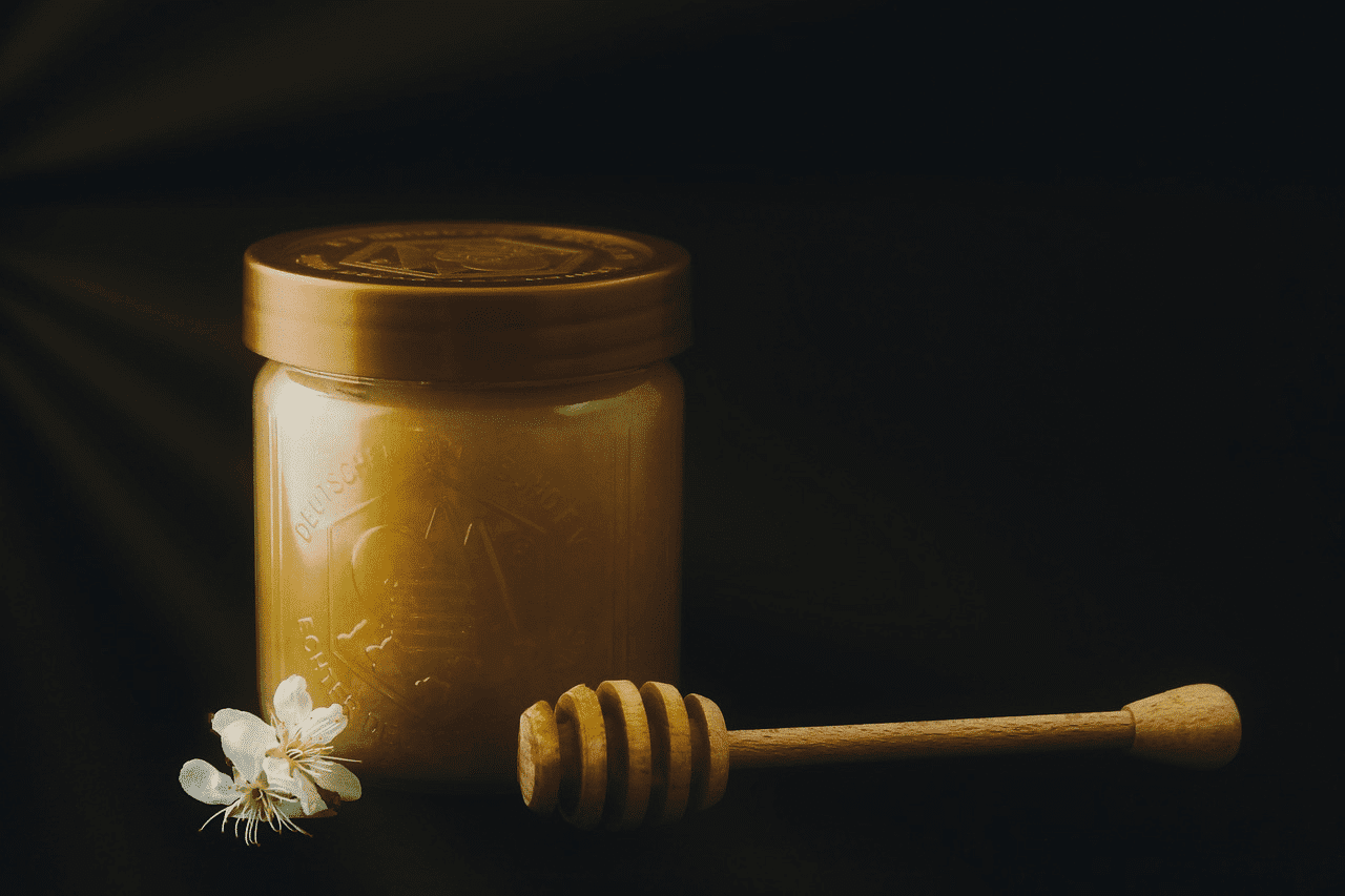 Jar of honey with a wooden dipper and white flower on a dark background.