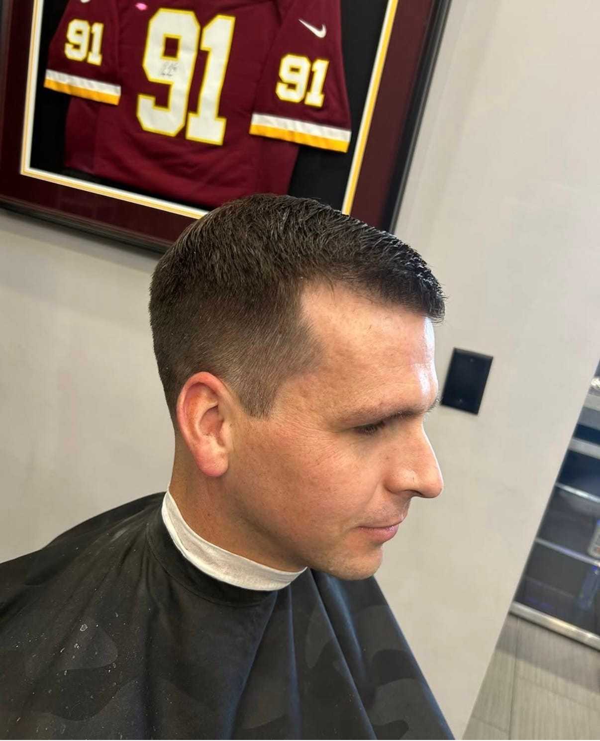 Man with short haircut in barber chair, sports jersey framed on wall.
