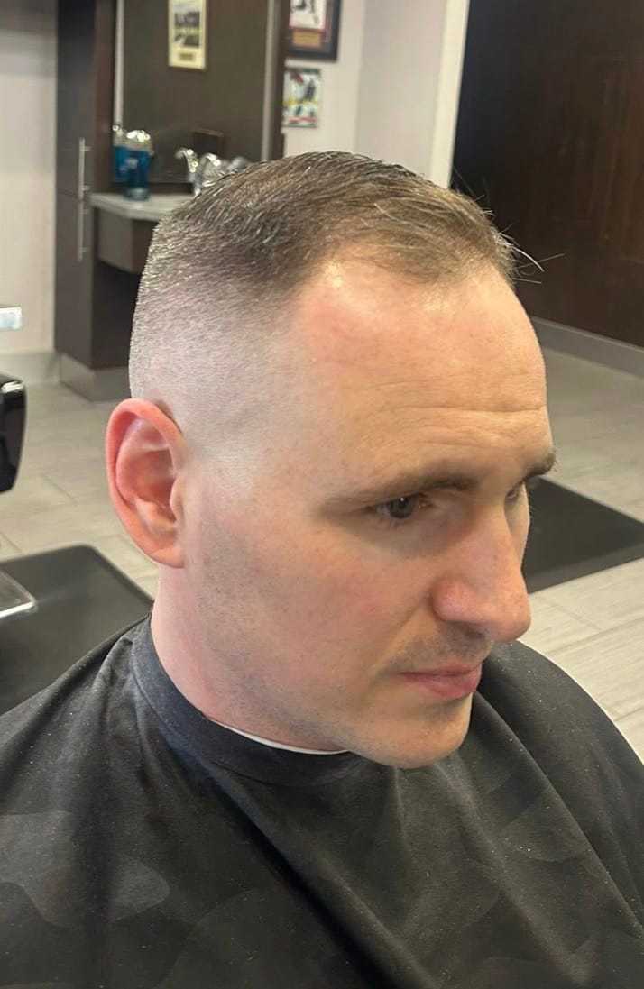 Man with a short haircut seated in a barber chair wearing a black cape.