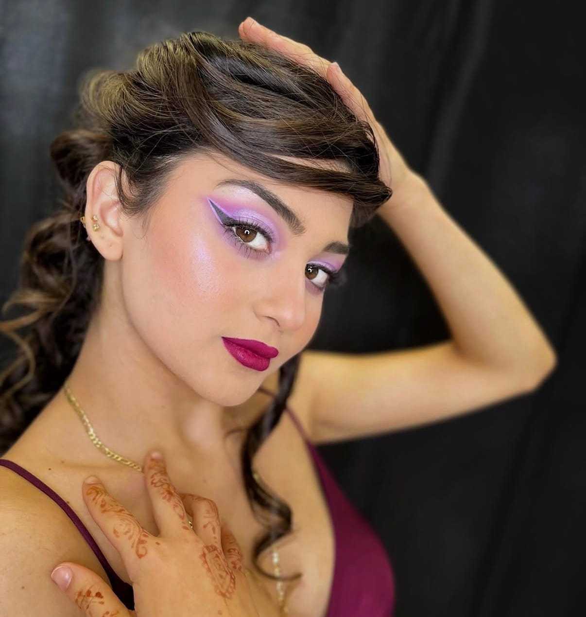 Woman with hennaed hand, purple eye makeup, and vibrant lipstick poses against dark background.
